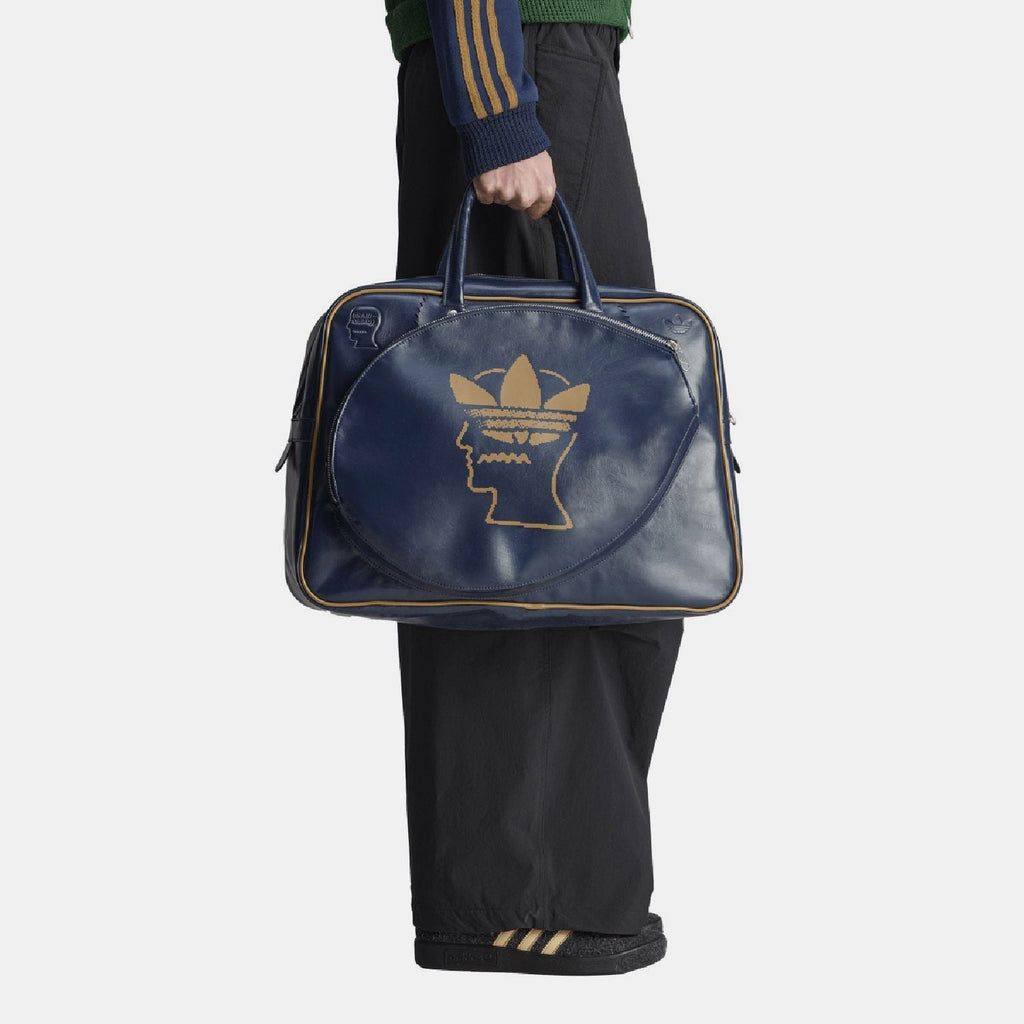 Navy duffel bag held in hand with front gold Trefoil head logo and side zipper ends.