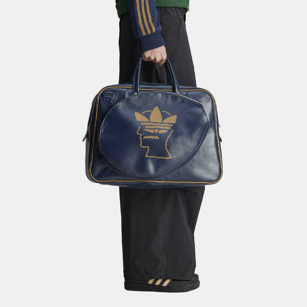 Navy duffel bag held in hand with front gold Trefoil head logo and side zipper ends.