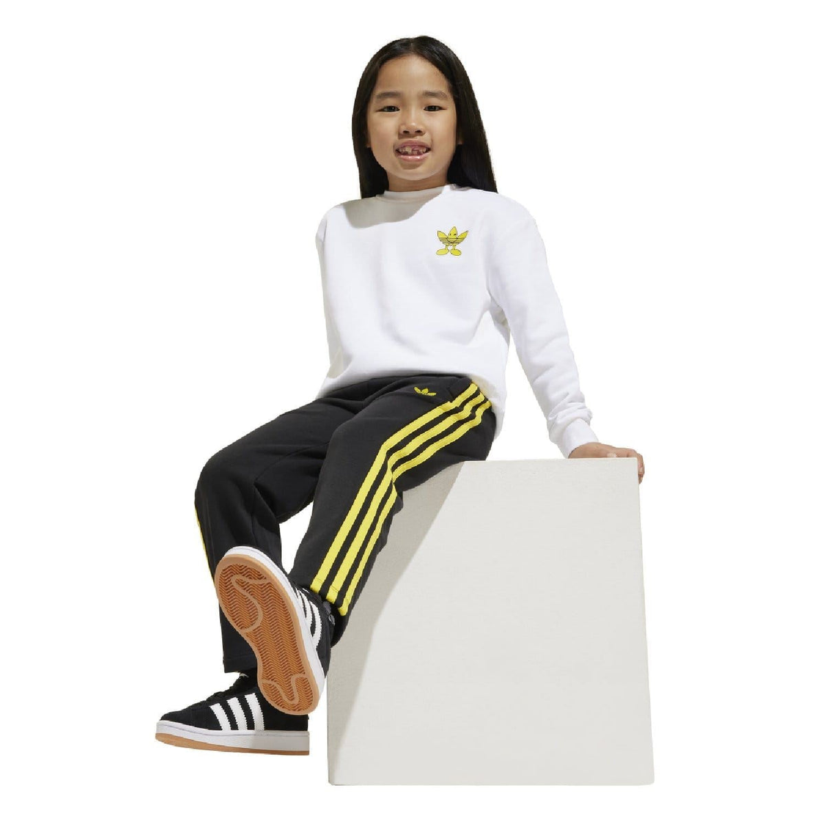 Kids white crew sweatshirt with yellow Smiley badge and black joggers with yellow side stripes.