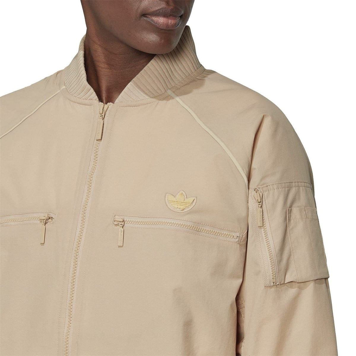 Beige bomber jacket close-up with funnel neck, zip front, and logo badge.