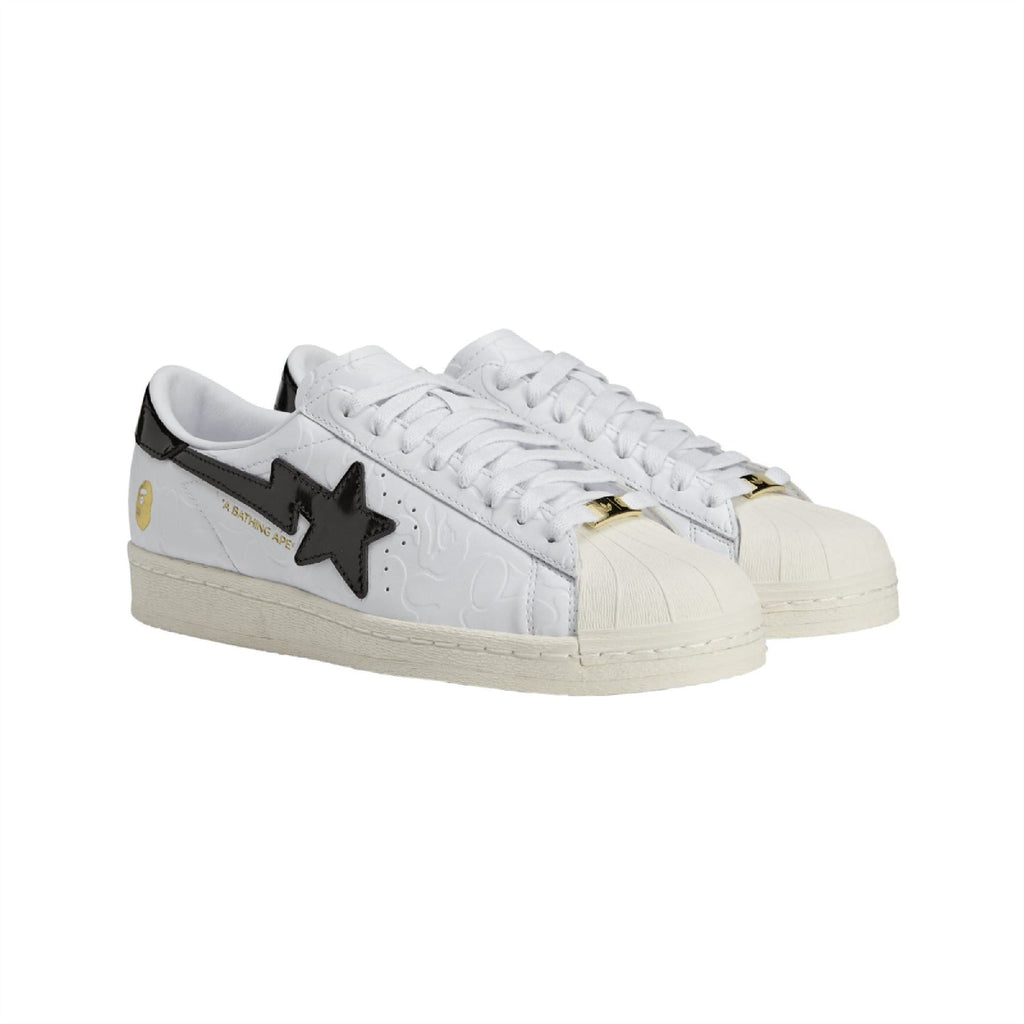 White low-top sneakers with black star emblem, gold lace hardware, and beige sole